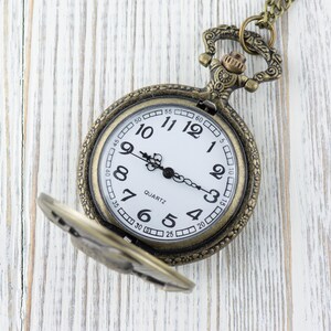 May include: A vintage-style pocket watch with a white face and black Roman numeral markings. The watch has a gold-toned metal case and a chain attached to the top.