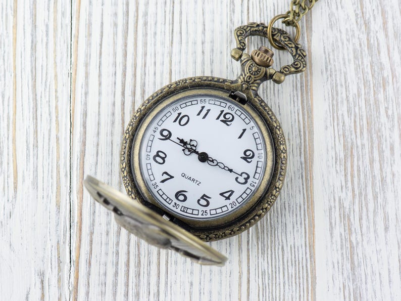 May include: A vintage-style pocket watch with a white face and black Roman numeral markings. The watch has a gold-toned metal case and a chain attached to the top.