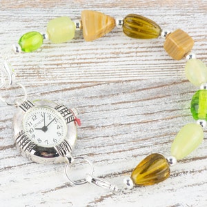 May include: A silver watch with a white face and black numbers is attached to a bracelet made of green, brown and beige beads.