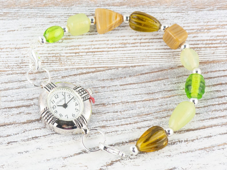 May include: A silver watch with a white face and black numbers is attached to a bracelet made of green, brown and beige beads.