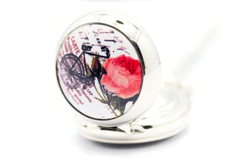 Puede incluir: Un medall&oacute;n plateado con fondo blanco y una imagen vintage de una bicicleta y una rosa rosa. El medall&oacute;n tiene una cadena plateada.