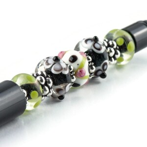 Op de afbeelding: Een zwarte pen met decoratieve glazen kralen. De kralen zijn wit, zwart en groen met bloemen- en stippenpatronen. Het ontwerp van de pen bevat zilverkleurige metalen accenten.