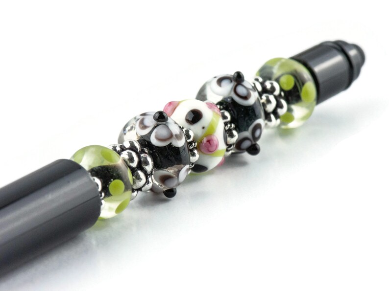Op de afbeelding: Een zwarte pen met decoratieve glazen kralen. De kralen zijn wit, zwart en groen met bloemen- en stippenpatronen. Het ontwerp van de pen bevat zilverkleurige metalen accenten.