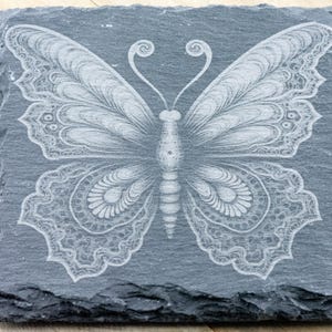 Puede incluir: Un posavasos de pizarra gris con un dise&ntilde;o de mariposa blanca grabada. La mariposa tiene detalles intrincados y un cuerpo en forma de coraz&oacute;n.