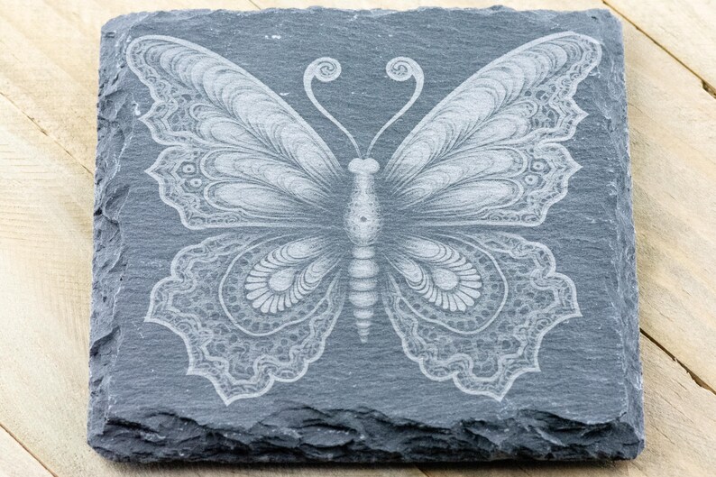 Puede incluir: Un posavasos de pizarra gris con un dise&ntilde;o de mariposa blanca grabada. La mariposa tiene detalles intrincados y un cuerpo en forma de coraz&oacute;n.