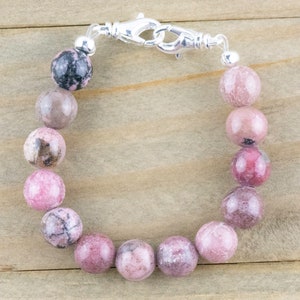 May include: A pink and gray gemstone bracelet with a silver clasp. The bracelet is made of round beads and is approximately 7 inches in length.
