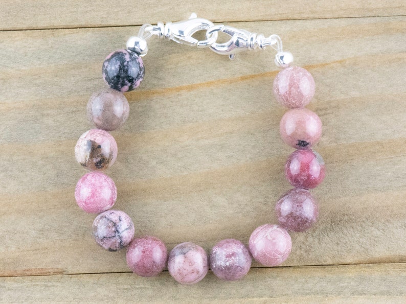 May include: A pink and gray gemstone bracelet with a silver clasp. The bracelet is made of round beads and is approximately 7 inches in length.