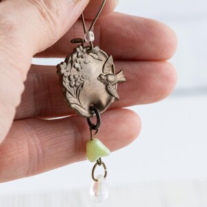 May include: A vintage-style pendant featuring an oval, bronze-colored charm with a bird and floral design. The pendant has a light green bead and a clear teardrop-shaped bead dangling from it. The jewelry is held up by a thin wire.