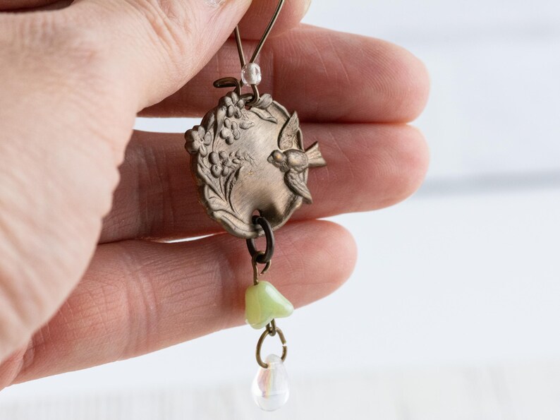 May include: A vintage-style pendant featuring an oval, bronze-colored charm with a bird and floral design. The pendant has a light green bead and a clear teardrop-shaped bead dangling from it. The jewelry is held up by a thin wire.