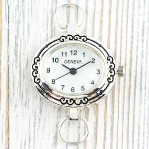 Può includere: Orologio ovale color argento con quadrante bianco e lancette nere. Il quadrante mostra la scritta "GENEVA" e numeri neri. L'orologio ha volute decorative attorno al quadrante e due anelli per il fissaggio.