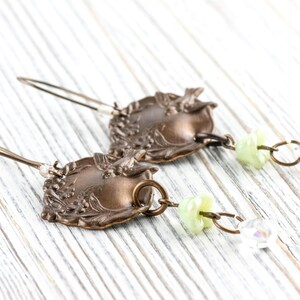 May include: A pair of vintage-style earrings. The earrings feature ornate, bronze-colored metalwork with floral and leaf designs. Each earring has a small, light green bead and a clear, iridescent crystal charm.