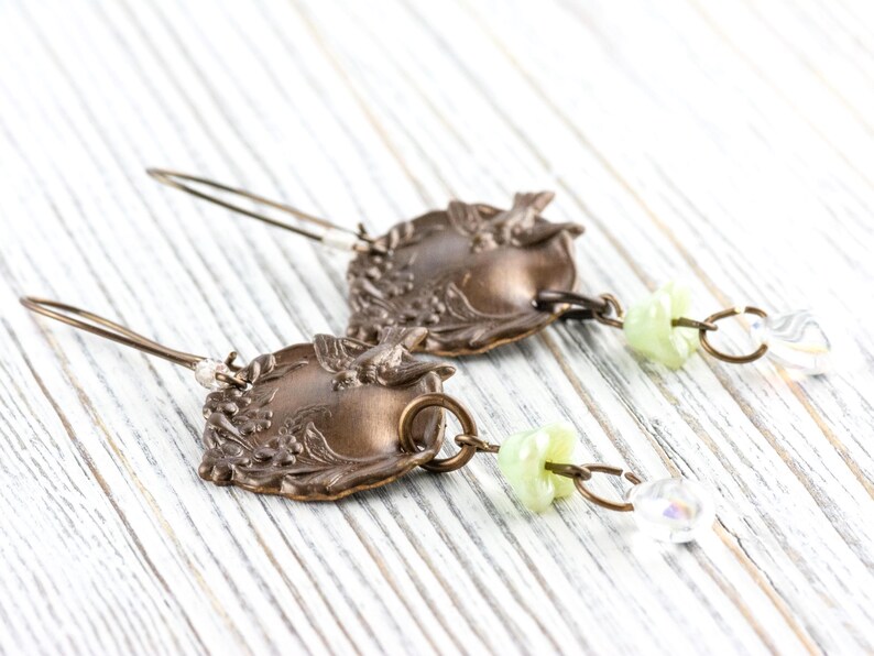 May include: A pair of vintage-style earrings. The earrings feature ornate, bronze-colored metalwork with floral and leaf designs. Each earring has a small, light green bead and a clear, iridescent crystal charm.