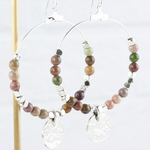 May include: Silver hoop earrings with a beaded design. The beads are a mix of brown, green, and pink colors. The earrings have a silver charm at the bottom.
