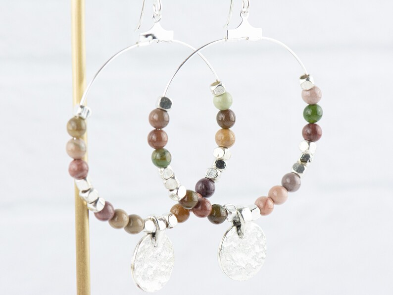 May include: Silver hoop earrings with a beaded design. The beads are a mix of brown, green, and pink colors. The earrings have a silver charm at the bottom.