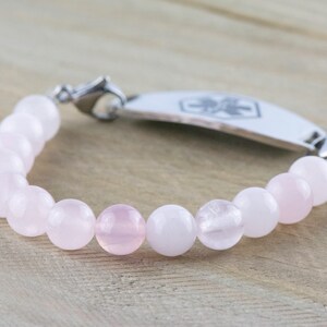 Puede incluir: Un brazalete de cuentas rosas y blancas con una placa de identificaci&oacute;n m&eacute;dica de plata. La placa tiene un s&iacute;mbolo de cruz roja.