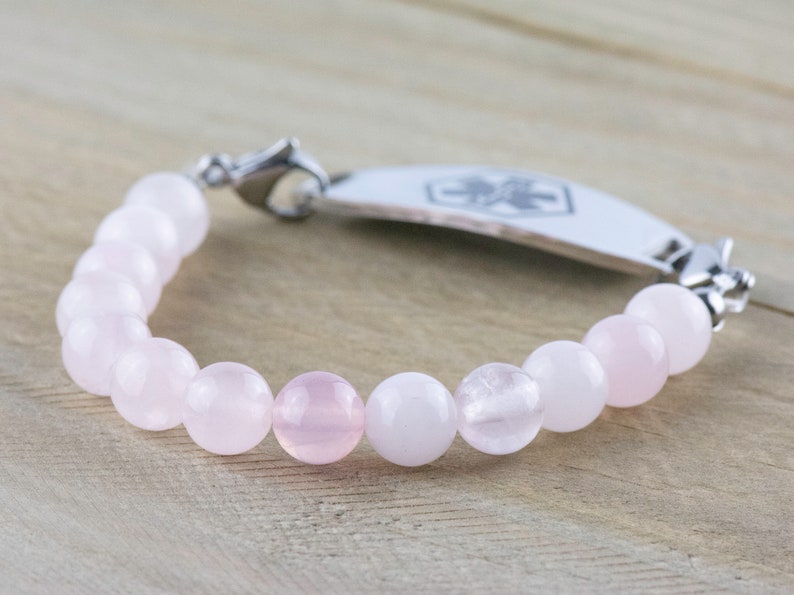 Puede incluir: Un brazalete de cuentas rosas y blancas con una placa de identificaci&oacute;n m&eacute;dica de plata. La placa tiene un s&iacute;mbolo de cruz roja.