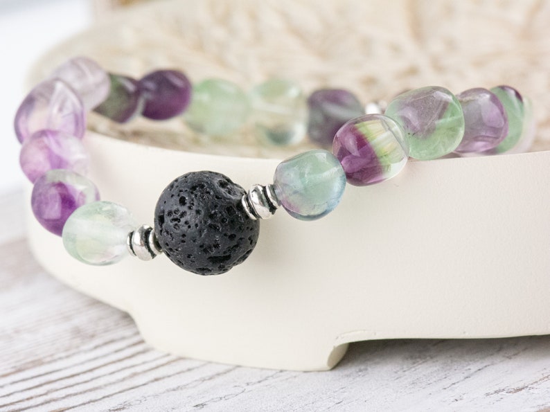 Puede incluir: Un brazalete hecho con cuentas de fluorita verde, p&uacute;rpura y blanca y una cuenta de lava negra. El brazalete est&aacute; sobre una superficie blanca.