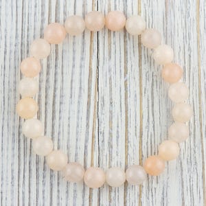Op de afbeelding: Een armband gemaakt van lichtroze en witte ovale kralen. De armband ligt op een wit houten oppervlak.