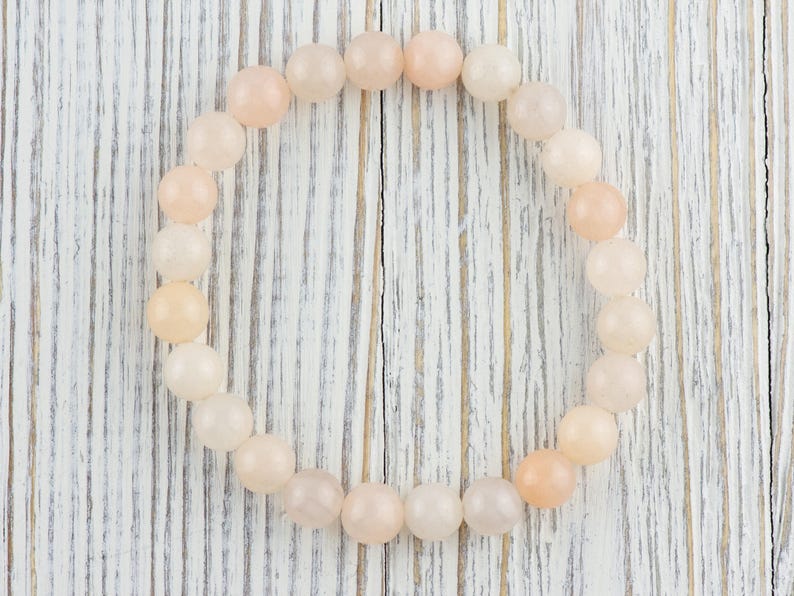 Op de afbeelding: Een armband gemaakt van lichtroze en witte ovale kralen. De armband ligt op een wit houten oppervlak.