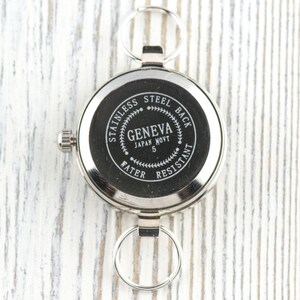 Pode incluir: Um rel&oacute;gio de bolso prateado com dois chaveiros. O mostrador do rel&oacute;gio &eacute; preto com texto branco que diz "STAINLESS STEEL BACK GENEVA JAPAN MOV'T 5 WATER RESISTANT". O rel&oacute;gio tem uma pequena coroa para dar corda.