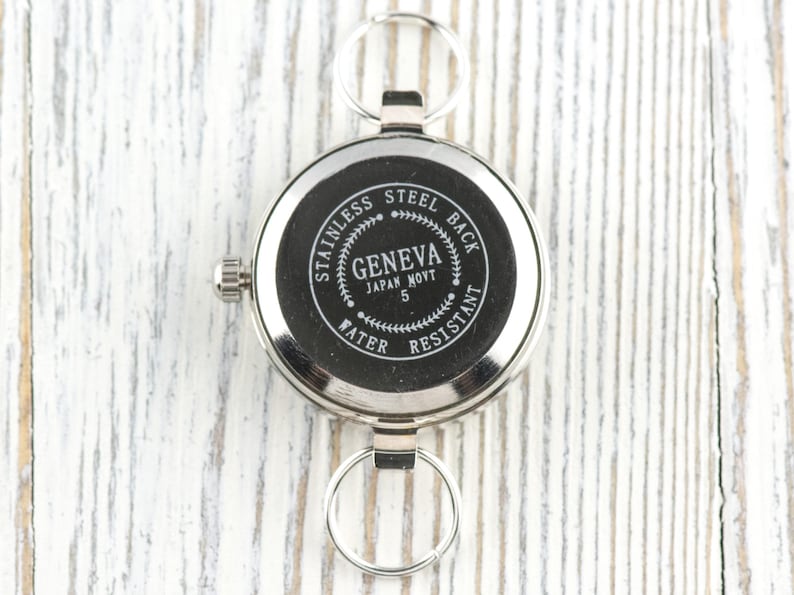 Pode incluir: Um rel&oacute;gio de bolso prateado com dois chaveiros. O mostrador do rel&oacute;gio &eacute; preto com texto branco que diz "STAINLESS STEEL BACK GENEVA JAPAN MOV'T 5 WATER RESISTANT". O rel&oacute;gio tem uma pequena coroa para dar corda.