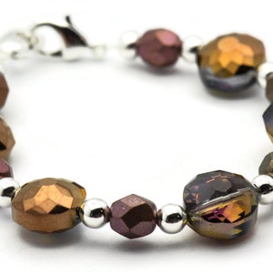 K&ouml;nnte beinhalten: Ein Armband aus facettierten Glasperlen in Braun-, Gold- und Lilat&ouml;nen. Die Perlen sind mit silbernen Metallperlen durchsetzt. Das Armband hat einen silbernen Verschluss.