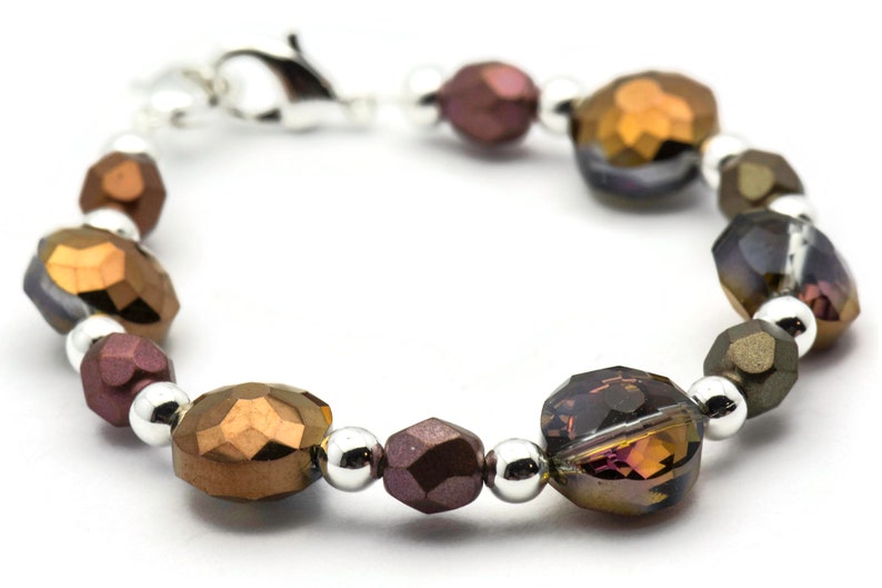 K&ouml;nnte beinhalten: Ein Armband aus facettierten Glasperlen in Braun-, Gold- und Lilat&ouml;nen. Die Perlen sind mit silbernen Metallperlen durchsetzt. Das Armband hat einen silbernen Verschluss.