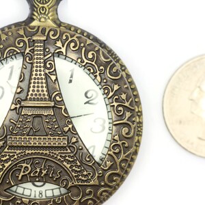 Może przedstawiać: Złoty zegarek kieszonkowy z ozdobnym wzorem przedstawiającym wieżę Eiffla i napis "Paris". Tarcza zegarka ma rzymskie cyfry godzinowe i białe tło.