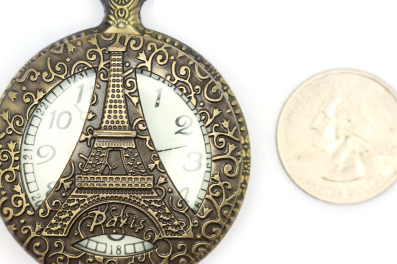 Może przedstawiać: Złoty zegarek kieszonkowy z ozdobnym wzorem przedstawiającym wieżę Eiffla i napis "Paris". Tarcza zegarka ma rzymskie cyfry godzinowe i białe tło.