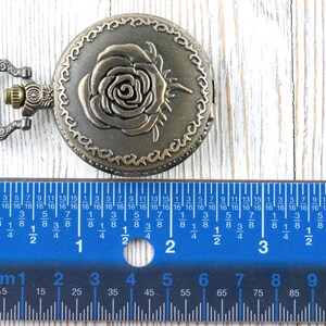 May include: Antique bronze-colored pocket watch with a rose design on the cover. The watch is round and has a decorative border. A blue ruler with inch and centimeter measurements is in the foreground.