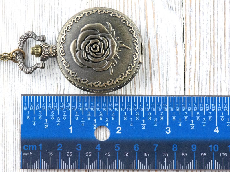 May include: Antique bronze-colored pocket watch with a rose design on the cover. The watch is round and has a decorative border. A blue ruler with inch and centimeter measurements is in the foreground.