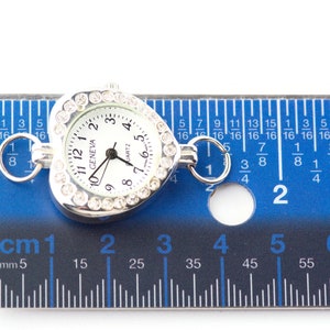 Pu&ograve; includere: Un orologio a forma di cuore argentato con quadrante bianco e dettagli in strass. L'orologio ha un diametro di 2,5 cm e un movimento al quarzo. L'orologio &egrave; mostrato accanto a un righello per riferimento alle dimensioni.
