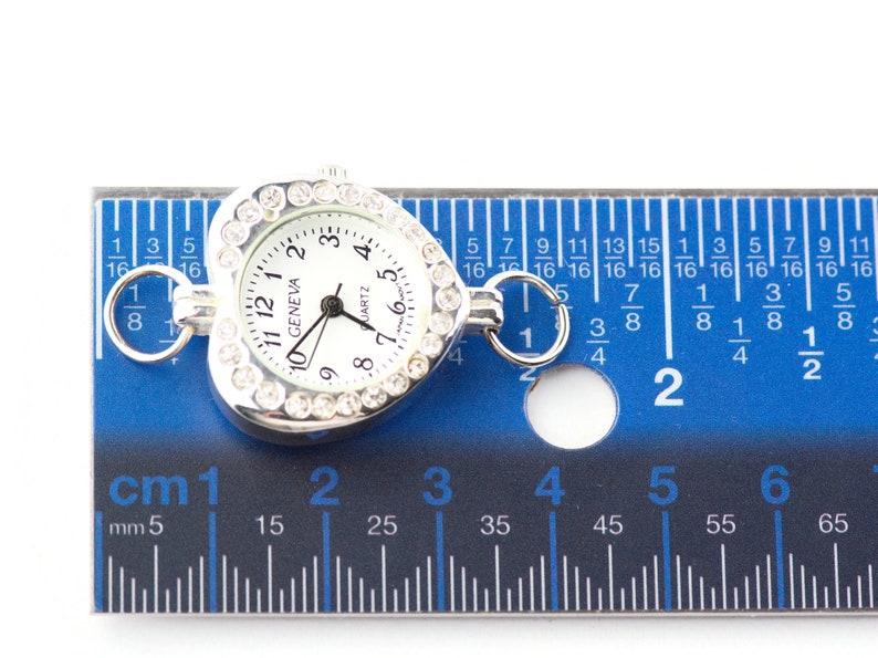 Pu&ograve; includere: Un orologio a forma di cuore argentato con quadrante bianco e dettagli in strass. L'orologio ha un diametro di 2,5 cm e un movimento al quarzo. L'orologio &egrave; mostrato accanto a un righello per riferimento alle dimensioni.