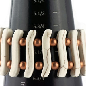 May include: White and copper beaded bracelet with a wavy design. The bracelet is shown on a black measuring tape with markings in inches.
