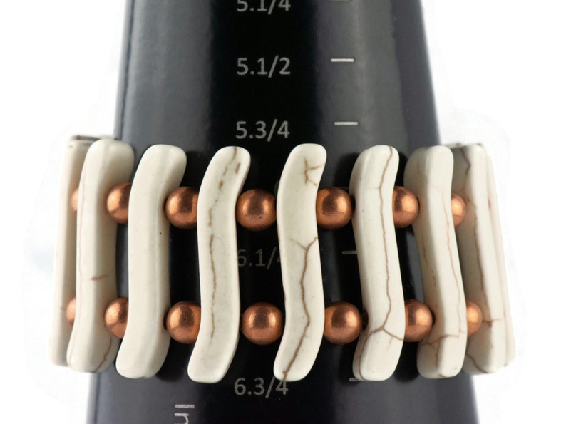 May include: White and copper beaded bracelet with a wavy design. The bracelet is shown on a black measuring tape with markings in inches.