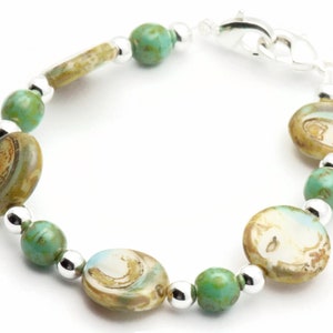 Op de afbeelding: Een turquoise en bruine glazen kralen armband met een zilveren sluiting. De armband heeft ovale kralen met een draaiende print.