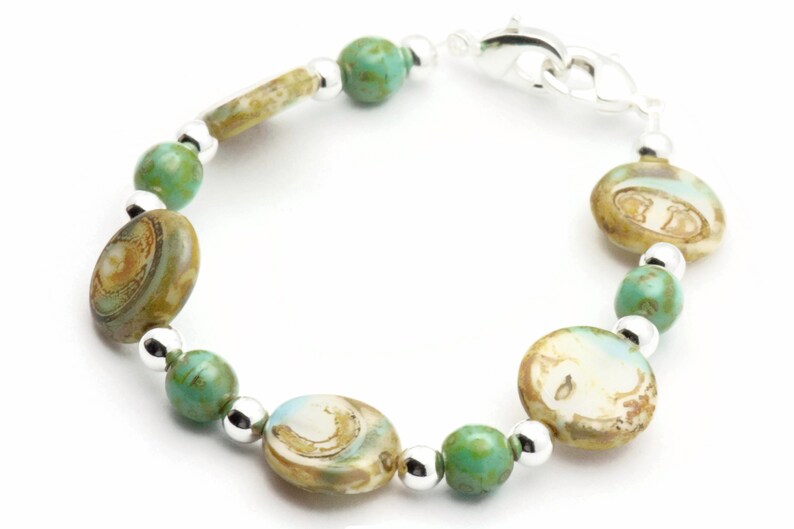 Op de afbeelding: Een turquoise en bruine glazen kralen armband met een zilveren sluiting. De armband heeft ovale kralen met een draaiende print.