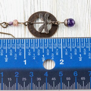 Może przedstawiać: Kolczyk w odcieniu brązu z zawieszką w kształcie ważki, fioletową koralikiem i małymi r&oacute;żowymi koralikami. Kolczyk ma około 5 cm długości, mierząc niebieską linijką na zdjęciu. Biżuteria.