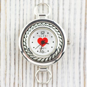 May include: Silver pocket watch with a white face. The watch face has a red heart, black dots, and the word "TRENDZ". It has a decorative rope-like border and two ring attachments. The time is set to 12:16.