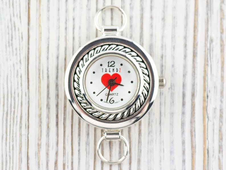 May include: Silver pocket watch with a white face. The watch face has a red heart, black dots, and the word "TRENDZ". It has a decorative rope-like border and two ring attachments. The time is set to 12:16.
