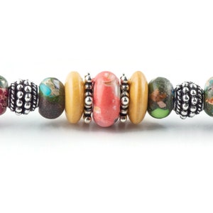 K&ouml;nnte beinhalten: Ein buntes Perlenarmband mit einer Mischung aus braunen, gr&uuml;nen, rosa und gelben Perlen. Das Armband hat silberne Akzente und schwarze Endkappen.