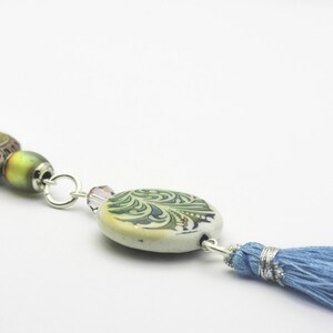 Puede incluir: Un collar de cadena plateada con cuentas verdes, marrones y moradas y un borla azul.