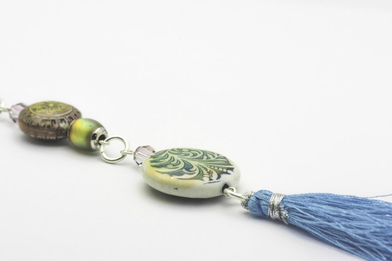 Puede incluir: Un collar de cadena plateada con cuentas verdes, marrones y moradas y un borla azul.
