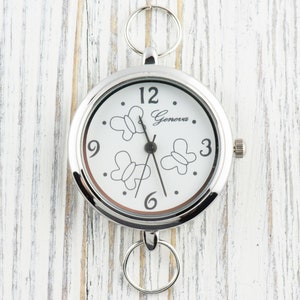Puede incluir: Un reloj de bolsillo redondo de tono plateado con esfera blanca que presenta contornos de mariposas negras y el texto Geneva. El reloj incluye manecillas negras de hora, minuto y segundo, n&uacute;meros negros en las posiciones 3, 6, 9 y 12, y dos anillos plateados unidos en la parte superior e inferior.