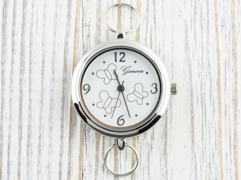 Puede incluir: Un reloj de bolsillo redondo de tono plateado con esfera blanca que presenta contornos de mariposas negras y el texto Geneva. El reloj incluye manecillas negras de hora, minuto y segundo, n&uacute;meros negros en las posiciones 3, 6, 9 y 12, y dos anillos plateados unidos en la parte superior e inferior.