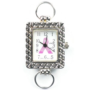 May include: A silver-toned rectangular pocket watch with a white face featuring black numerals and a pink ribbon. The watch has a textured frame and two circular attachments. The watch face reads "GENEVA QUARTZ".