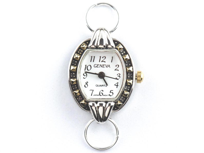 Peut inclure: Montre Geneva &agrave; quartz ovale avec un cadran blanc, des aiguilles noires et des accents dor&eacute;s. La montre a une lunette argent&eacute;e avec des clous noirs et dor&eacute;s. La montre est attach&eacute;e &agrave; un anneau argent&eacute; en haut et en bas.