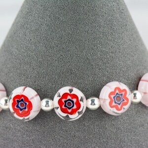 Puede incluir: Un brazalete con cuentas de vidrio rosa y blanco con dise&ntilde;os florales rojos y azules. Las cuentas est&aacute;n separadas por cuentas espaciadoras plateadas.
