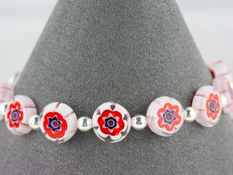 Puede incluir: Un brazalete con cuentas de vidrio rosa y blanco con dise&ntilde;os florales rojos y azules. Las cuentas est&aacute;n separadas por cuentas espaciadoras plateadas.