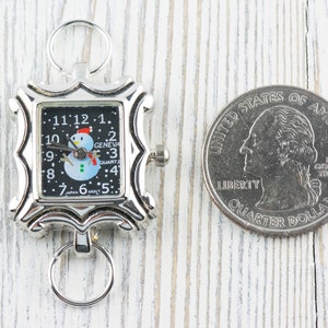 May include: A silver-toned pocket watch with a black face and a snowman design. The watch is about the size of a US quarter.